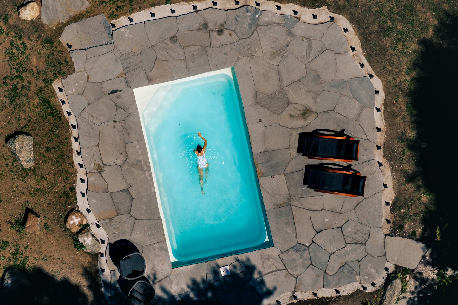 Une personne nage dans une piscine rectangulaire entourée d'une terrasse en pierre et de chaises longues.