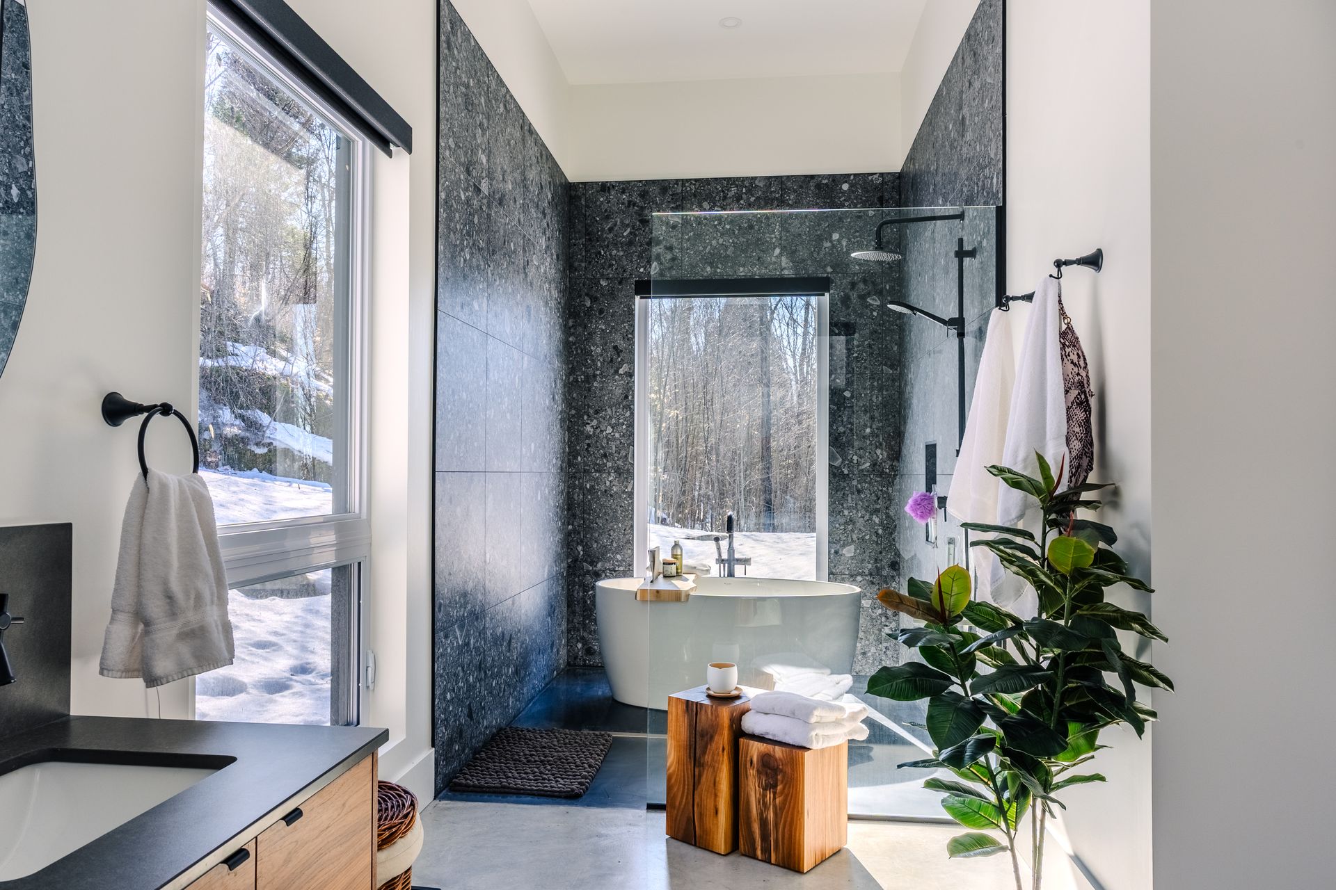 Salle de bains moderne avec carrelage en mosaïque noire, baignoire îlot et vue sur des arbres enneigés.