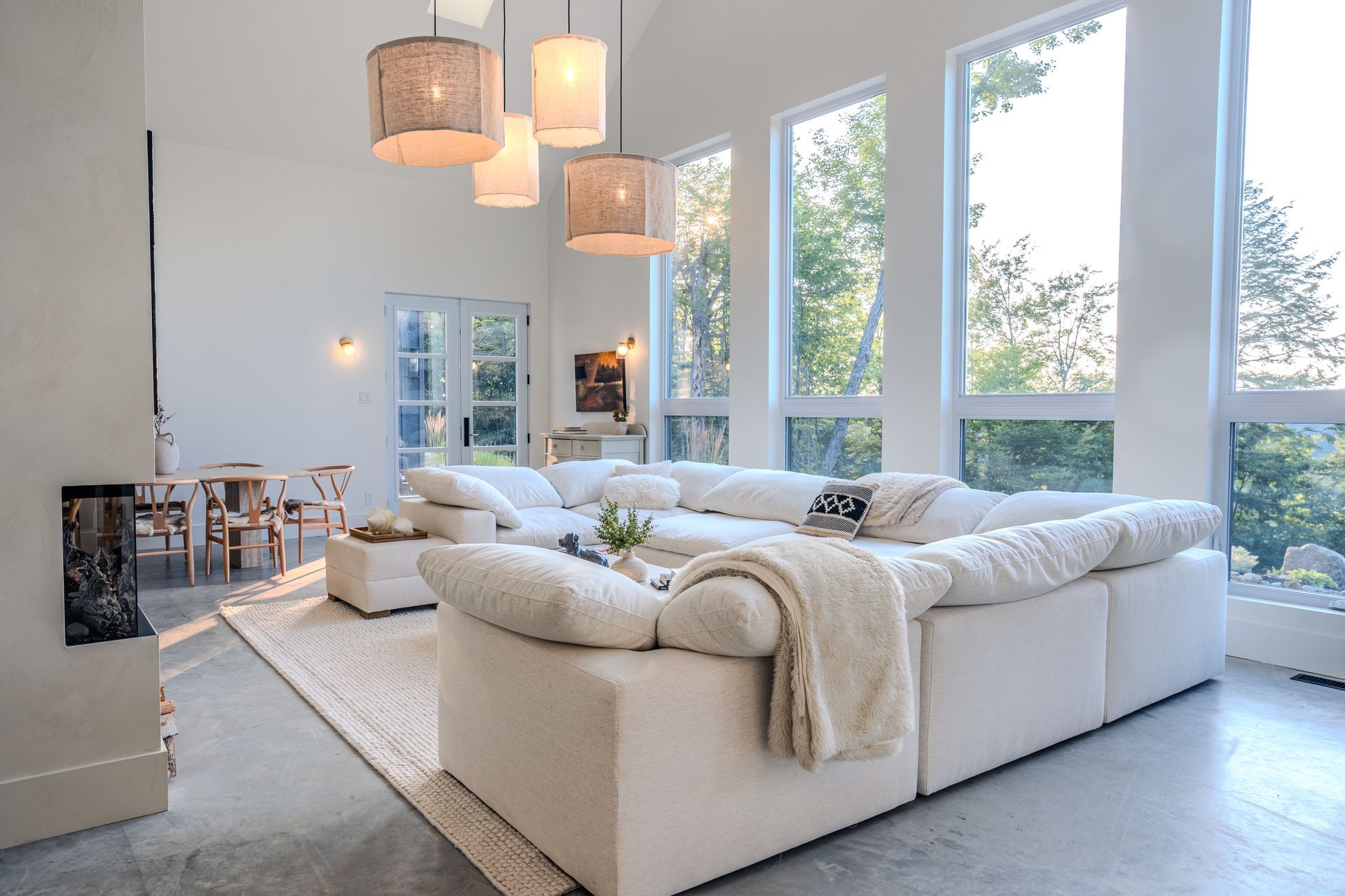 Salon lumineux et moderne avec un grand canapé d'angle blanc, de hautes fenêtres et des suspensions lumineuses.