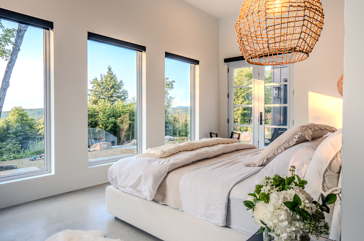 Chambre aux murs blancs, grandes fenêtres donnant sur un paysage verdoyant, lit avec draps blancs et suspension en osier.