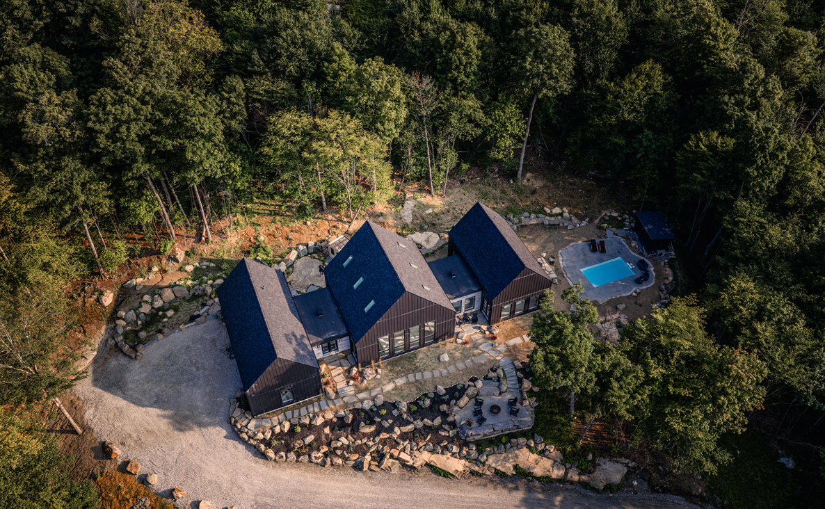 Vue aérienne d'un chalet moderne avec un toit sombre et une petite piscine entourée d'arbres.