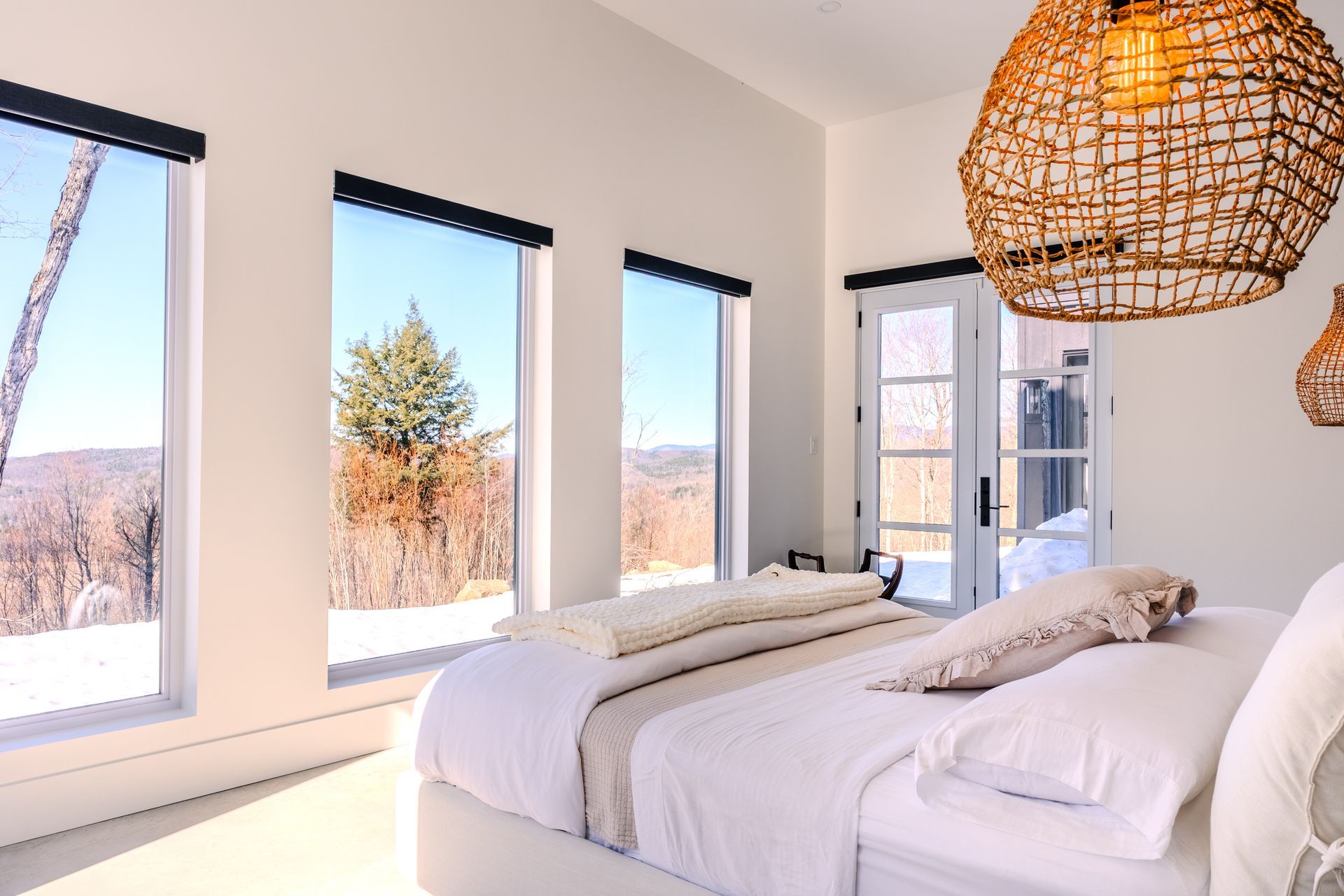 Chambre lumineuse avec un lit, de grandes fenêtres et un luminaire en osier. Vue sur un paysage enneigé.