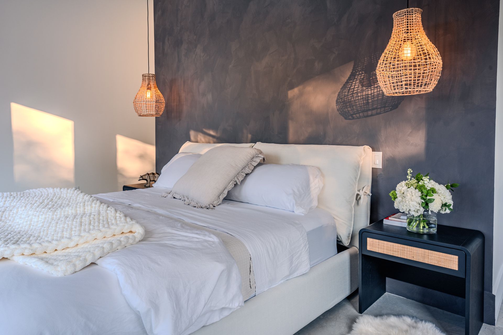 Chambre avec literie blanche, mur d'accent gris foncé et suspensions lumineuses en fibres tressées.