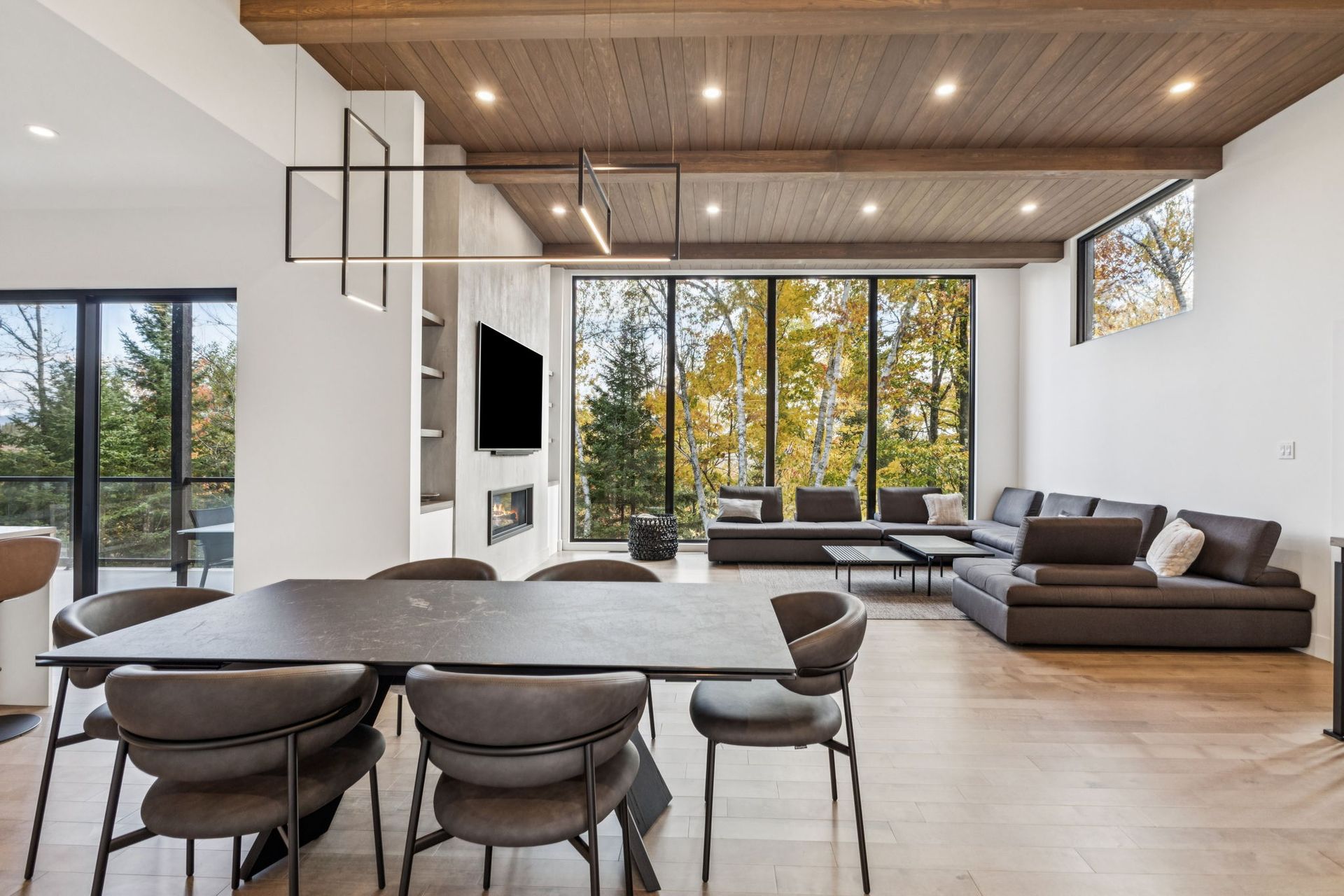 Salon moderne avec coin repas, grandes fenêtres donnant sur les arbres, poutres apparentes au plafond et canapé d'angle.