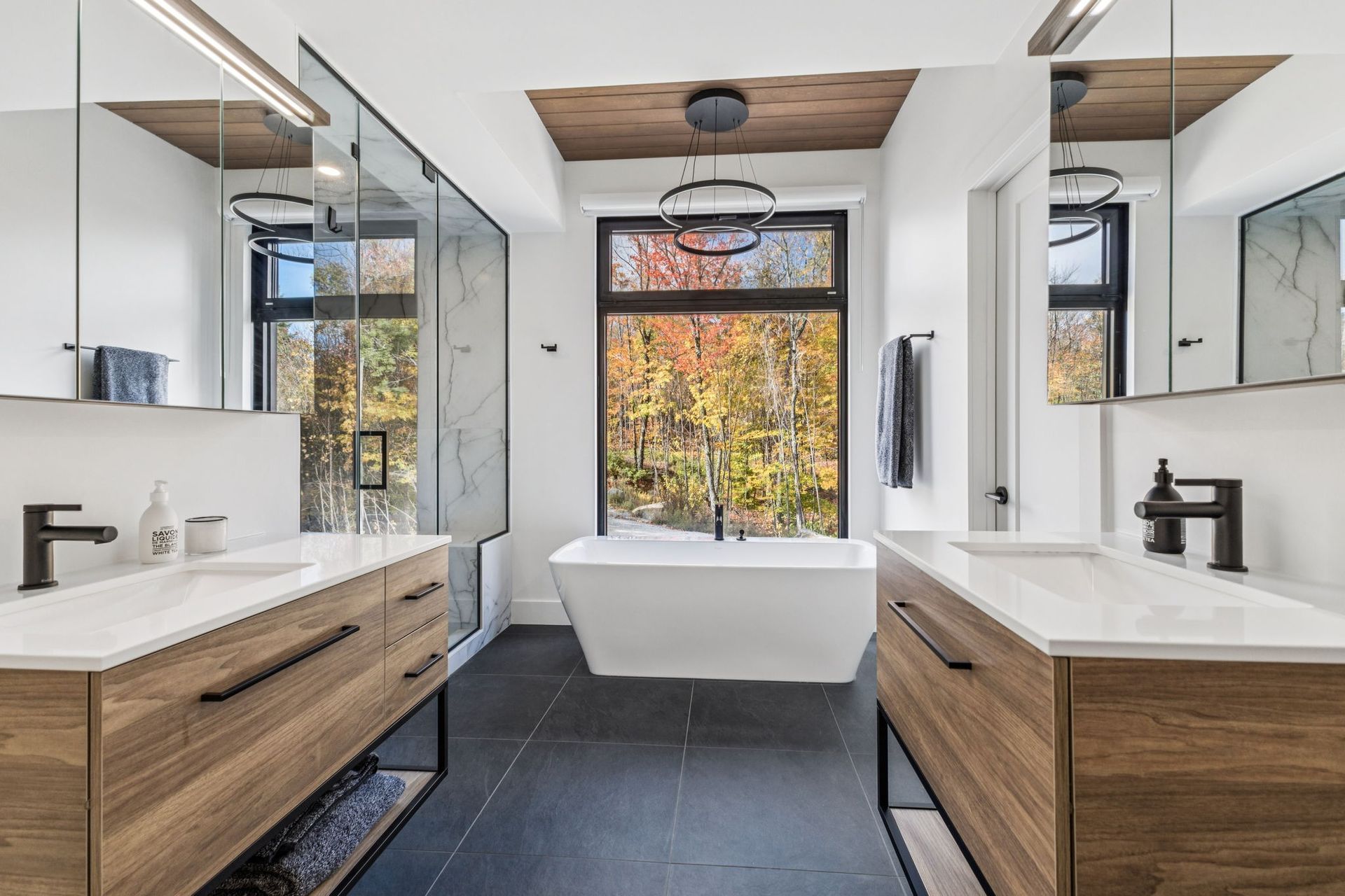 Salle de bains moderne avec baignoire blanche, meubles-lavabos en bois et grande fenêtre donnant sur le feuillage d'automne.