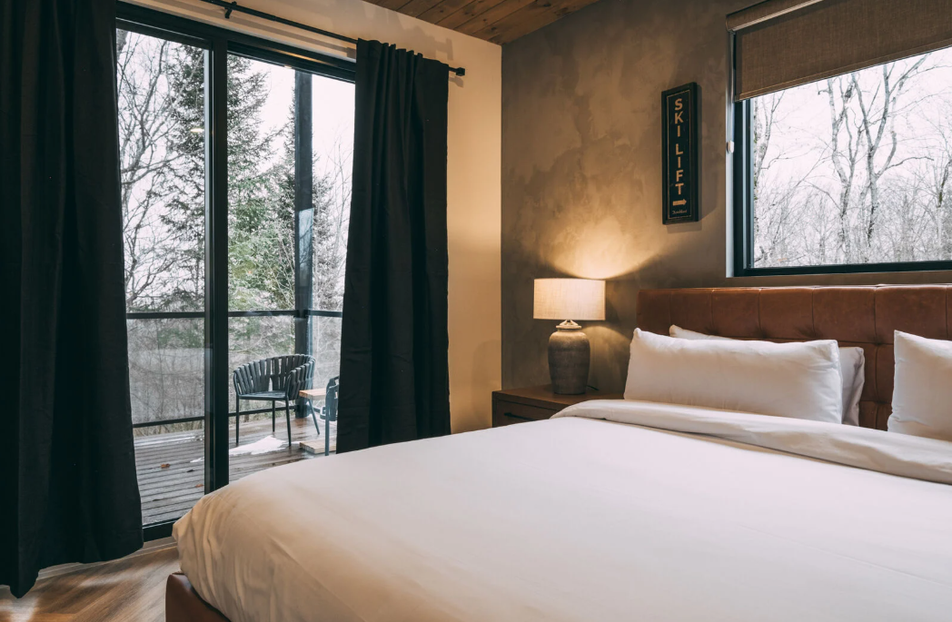 Chambre avec un grand lit, des rideaux sombres et une vue sur les arbres à travers une porte coulissante en verre.