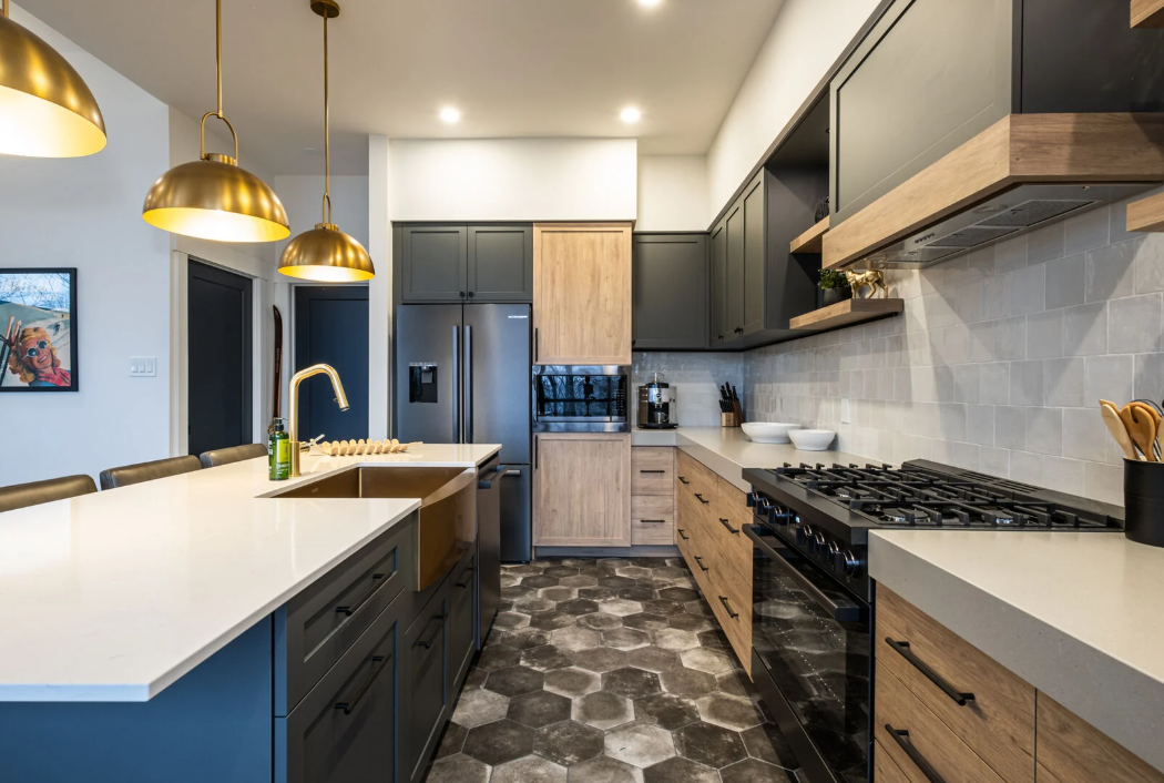 Cuisine moderne avec suspensions lumineuses dorées, armoires noires, éléments en bois et sol en carrelage géométrique.