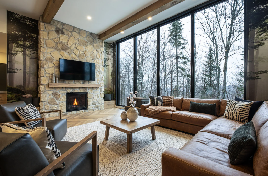 Salon avec cheminée en pierre, grande fenêtre donnant sur les arbres, canapé d'angle en cuir marron et éléments en bois.