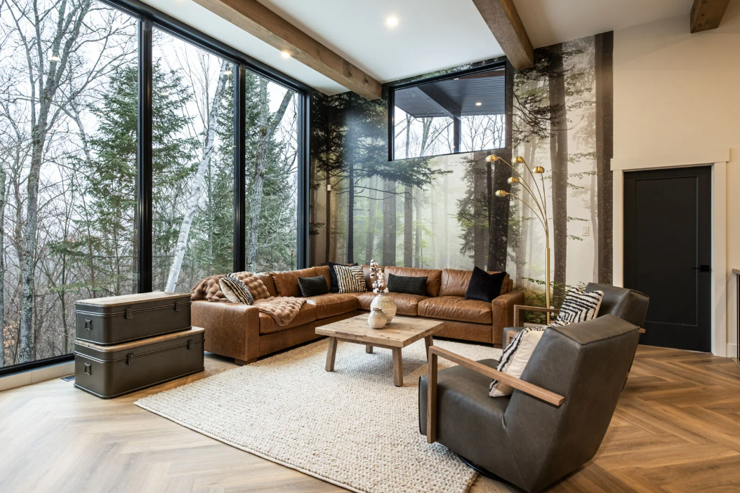 Salon avec de grandes fenêtres donnant sur les arbres, canapé d'angle en cuir, tapis et fauteuils d'appoint.