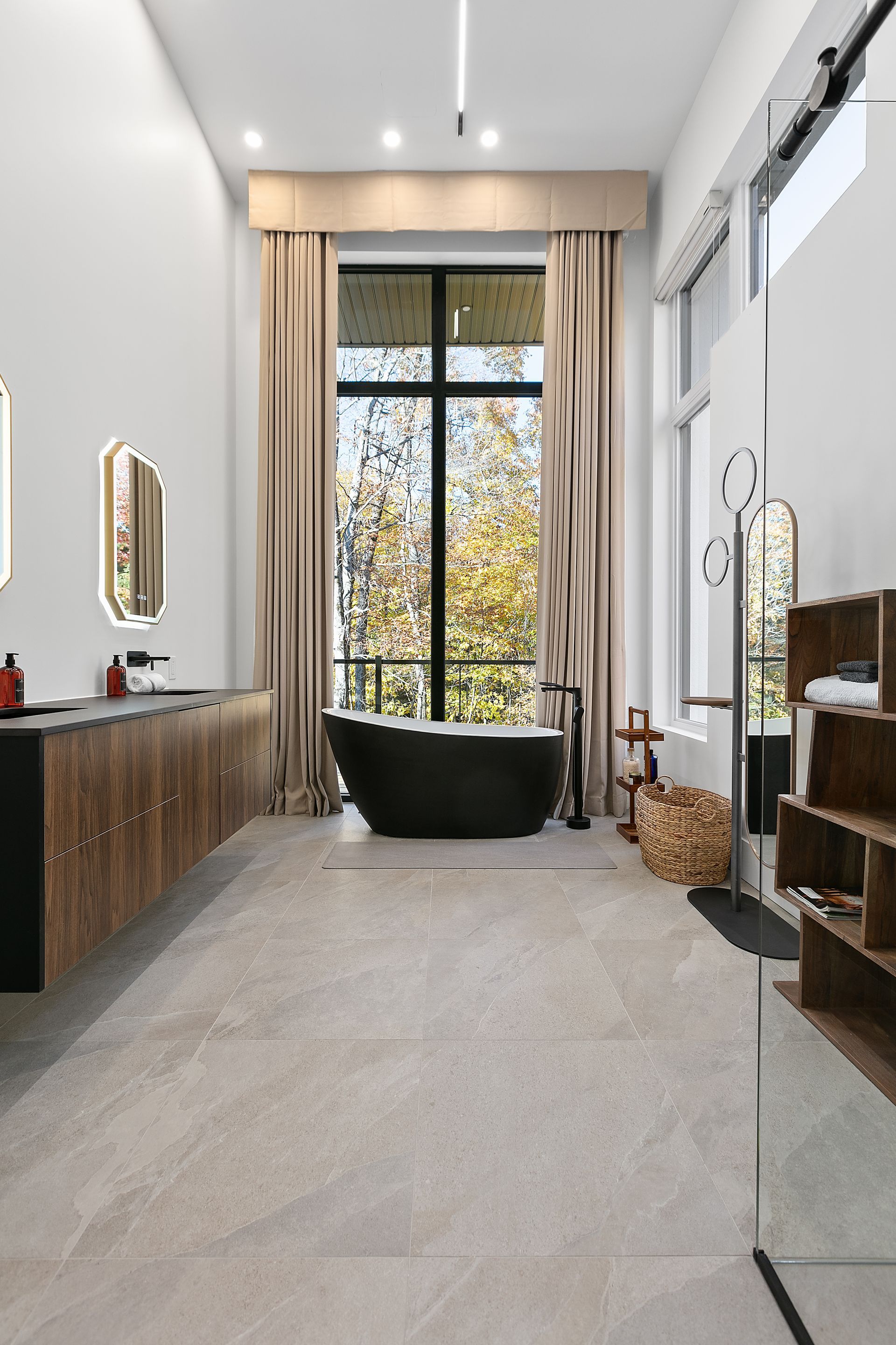 Salle de bains moderne avec baignoire noire devant une grande fenêtre ; meuble-lavabo en bois et sol beige clair.