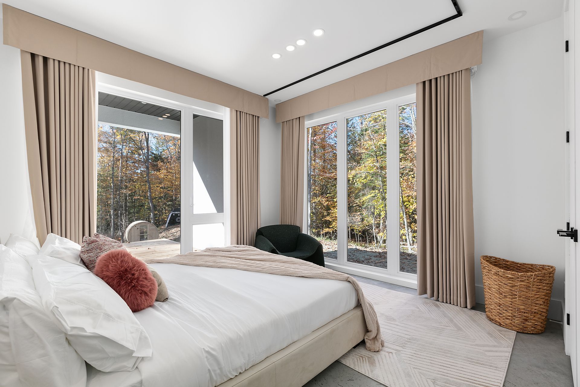 Chambre avec lit, grandes fenêtres et rideaux aux tons neutres. Un panier en osier est posé à côté du lit.