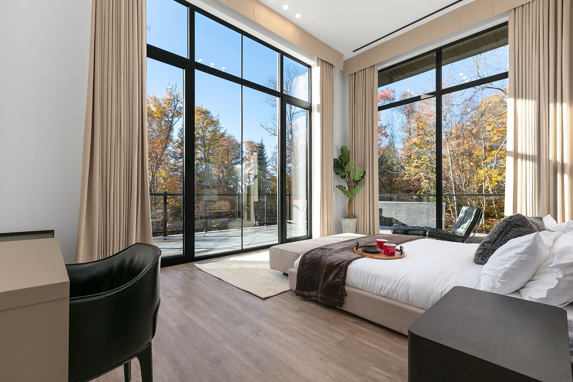 Chambre avec de grandes fenêtres donnant sur les arbres ; rideaux beiges, lit blanc, fauteuil foncé, parquet.