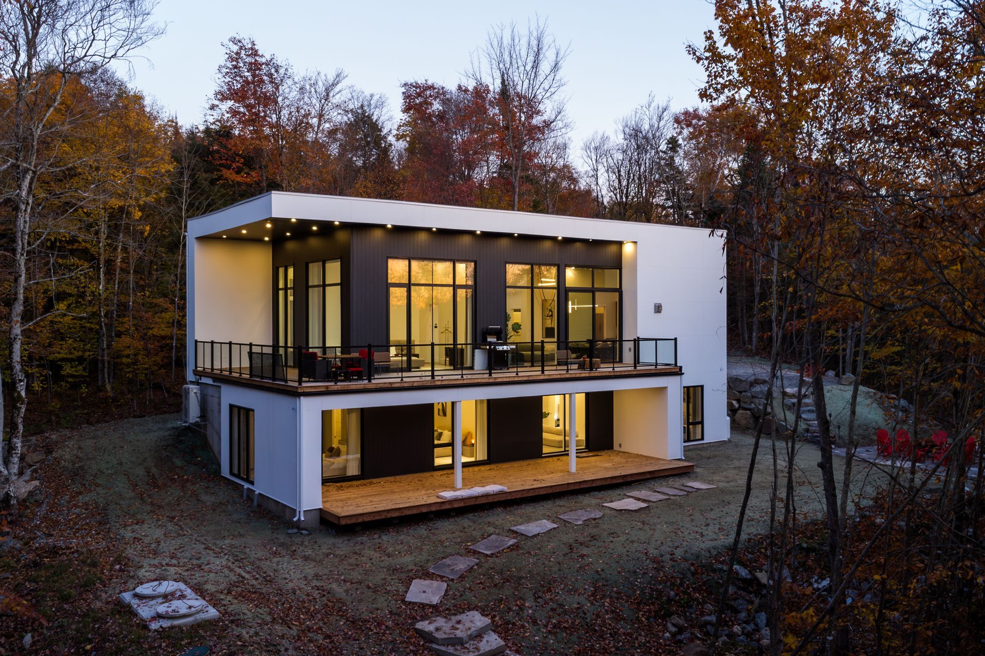 Maison moderne à deux étages avec fenêtres à cadre noir, murs blancs et terrasse en bois, entourée d'arbres aux couleurs d'automne.