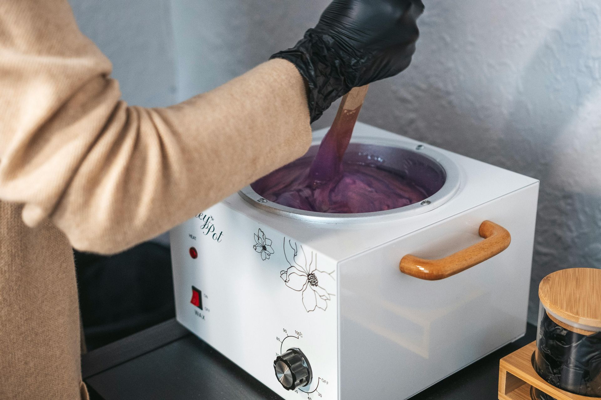Person stirring purple wax in a white wax warmer with a wooden handle; black gloved hand.