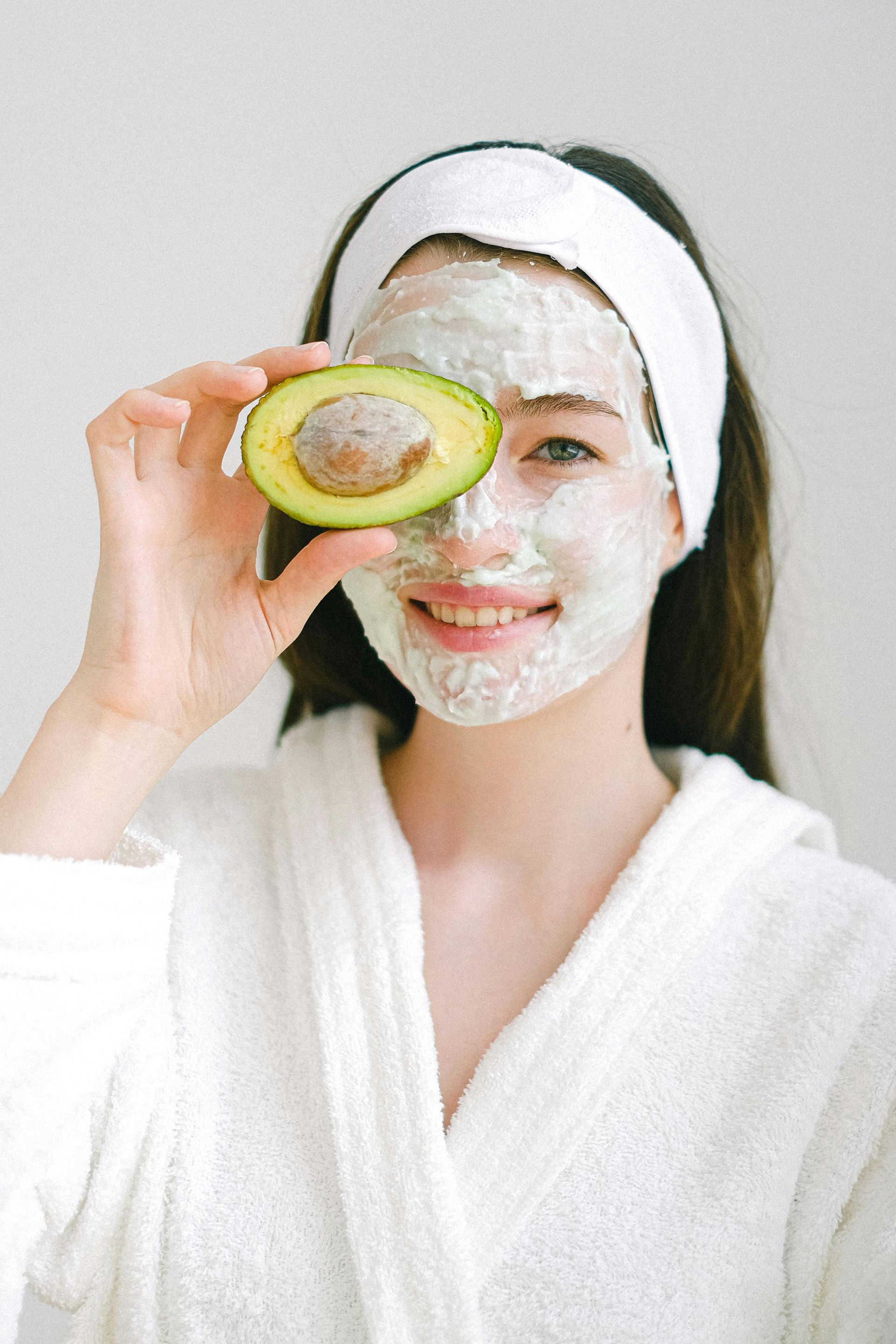 Woman with a face mask and avocado slice covering one eye, wearing a bathrobe and headband, smiling.