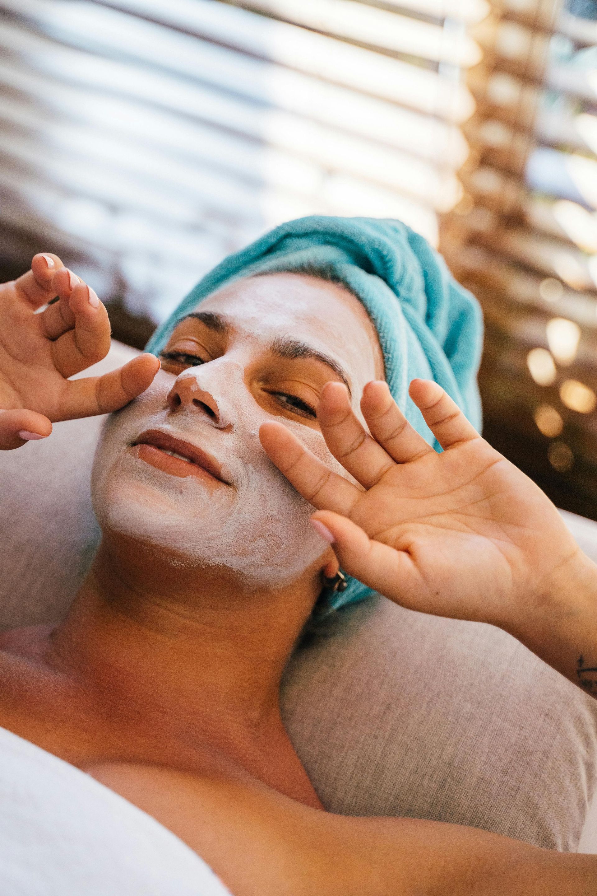 Person with facial mask, relaxing with hands near face, wearing a blue towel.