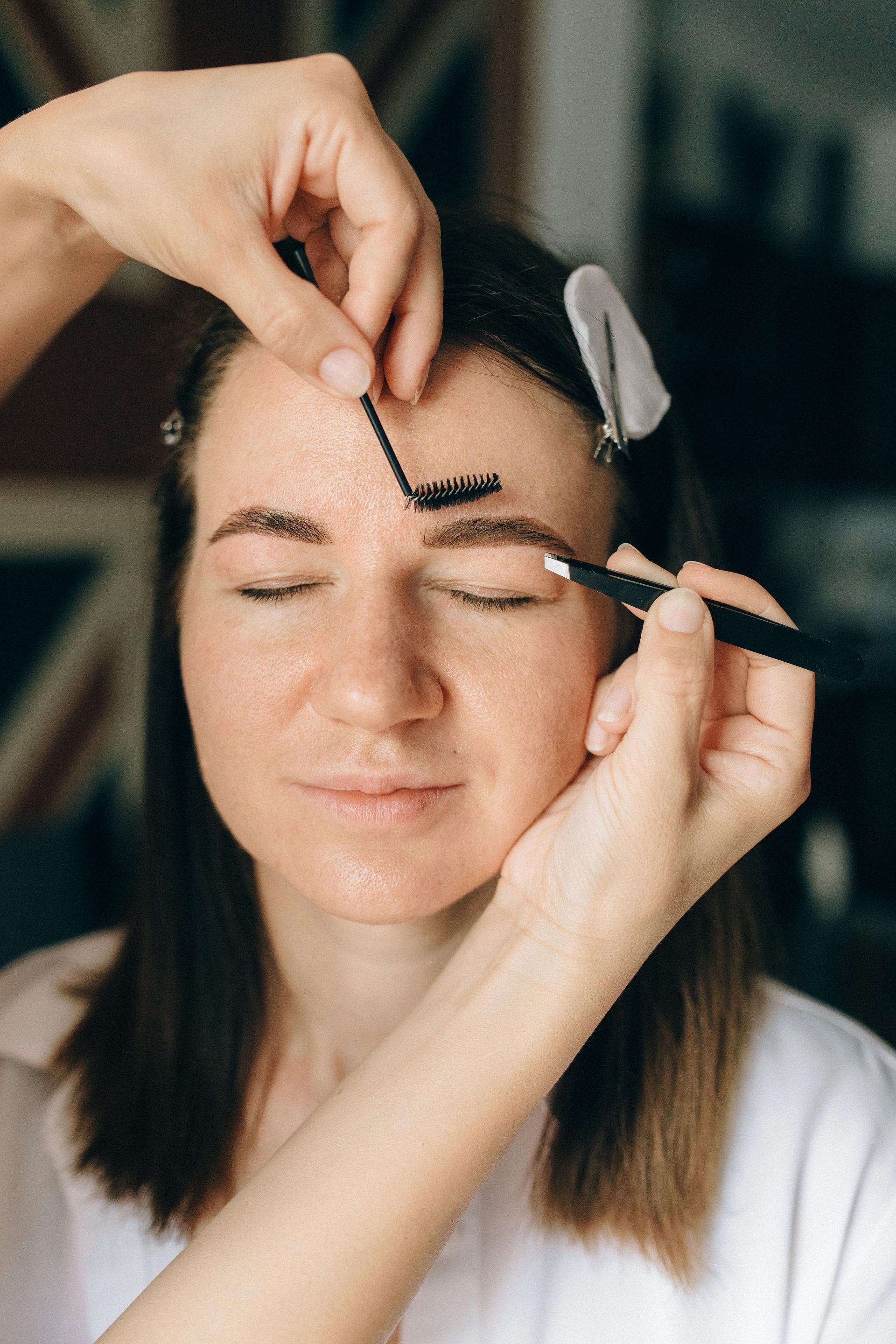 Person's eyebrows being styled by two hands, using comb and brush tools. Interior setting.