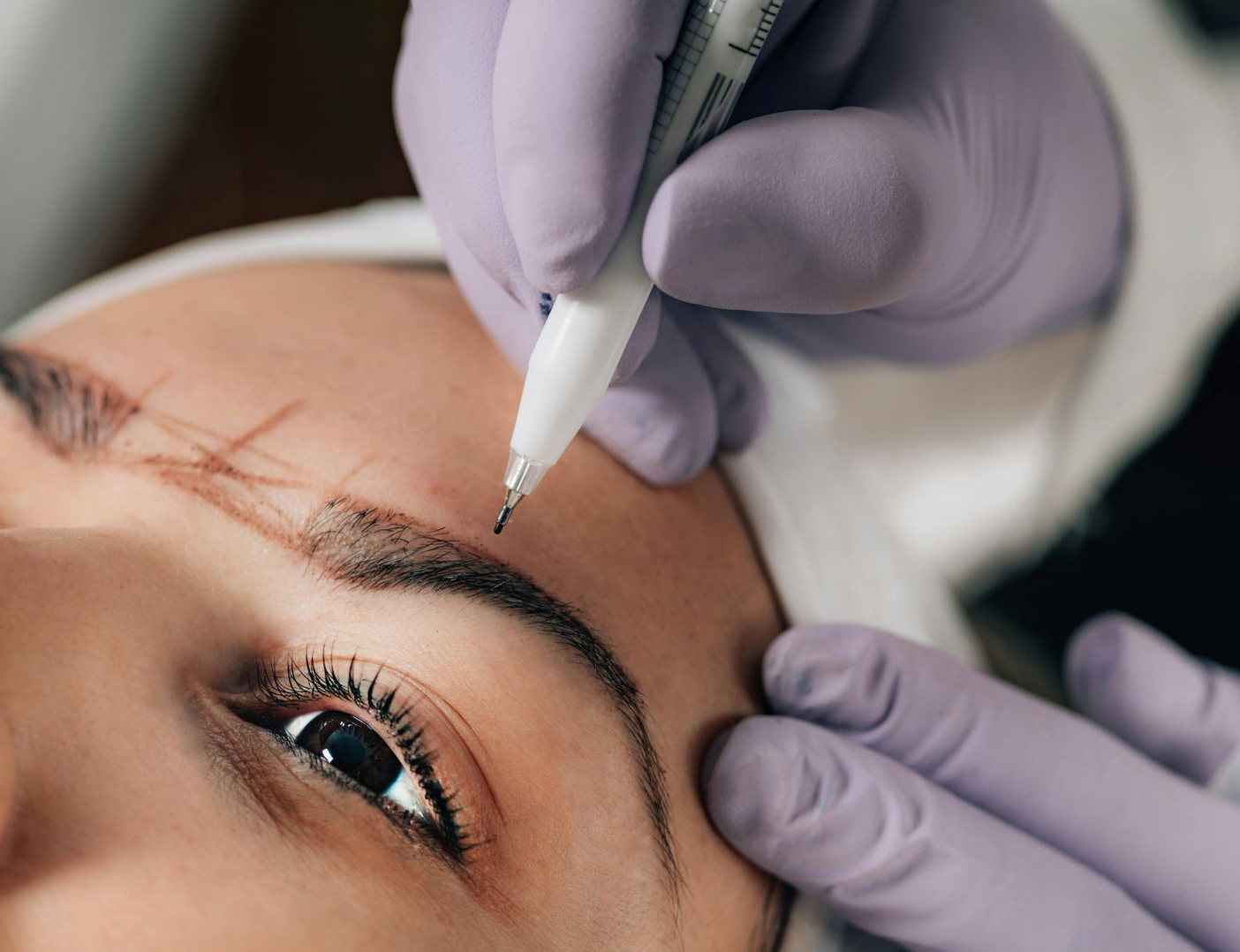 A person receiving microblading; technician's gloved hands using a pen to draw on eyebrow shape.