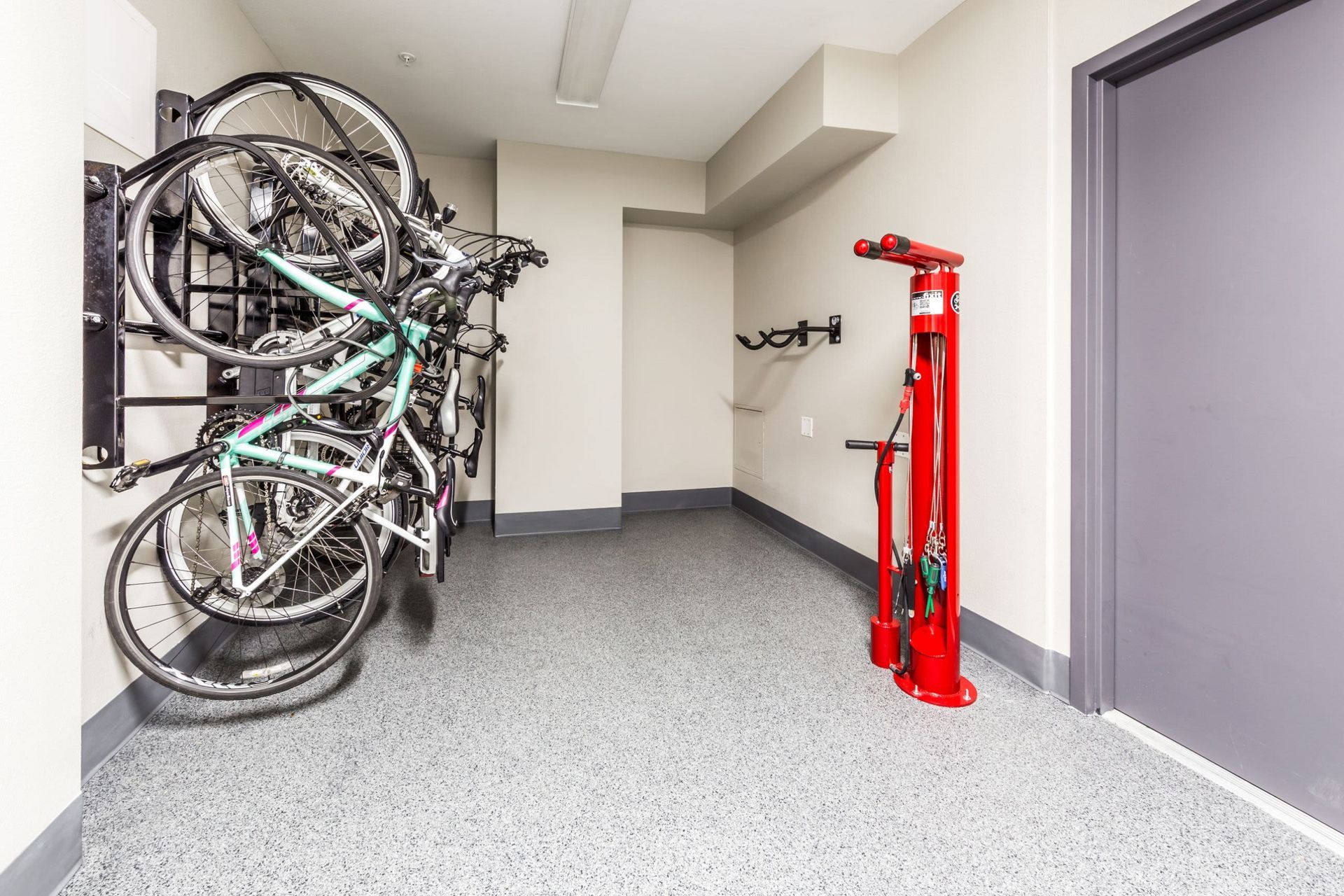A clean and organized indoor bicycle storage room with multiple bikes mounted on a vertical bike rack on the left wall. To the right stands a tall red manual bike pump, and hooks for hanging bike accessories are visible on the far wall. The room has a speckled grey floor, white walls, and fluorescent lighting, creating a bright and functional space for cyclists at Marq 211 in Seattle, WA.