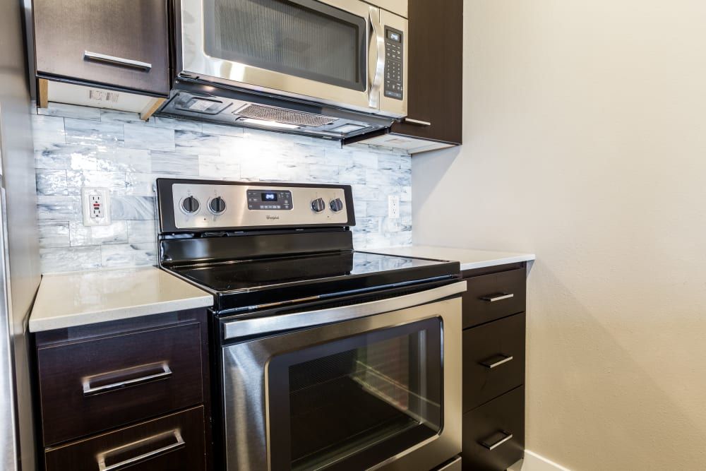 Modern kitchen with dark wood cabinets, stainless steel appliances including a range with an oven and a microwave above it, set against a backsplash with light gray and white marble-like tiles at Marq 211 in Seattle, WA.