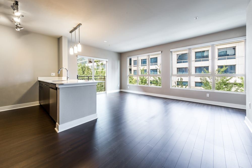 Modern empty apartment interior with large windows, dark hardwood floors, and a simple kitchen island with sink and pendant lighting. The room is bright, with natural light filtering in through the windows, which have a view of green trees and a neighboring building at Marq 211 in Seattle, WA.