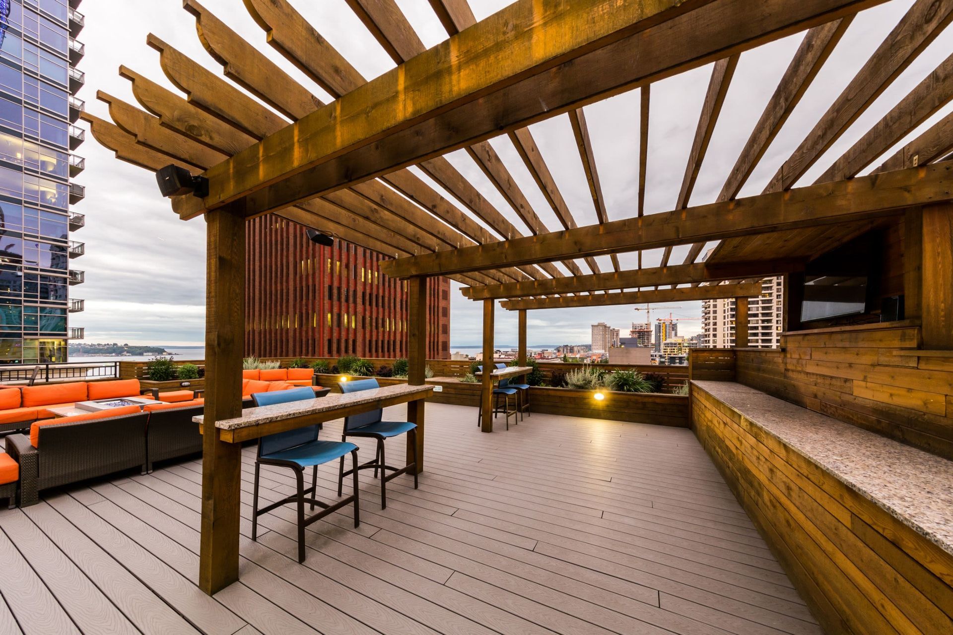 A modern rooftop terrace with wooden pergolas, featuring comfortable seating with orange cushions, high stools at a bar table, and an urban skyline view in the background. The space is inviting with warm lighting accents and a view of highrise buildings under a cloudy sky at Marq 211 in Seattle, WA.