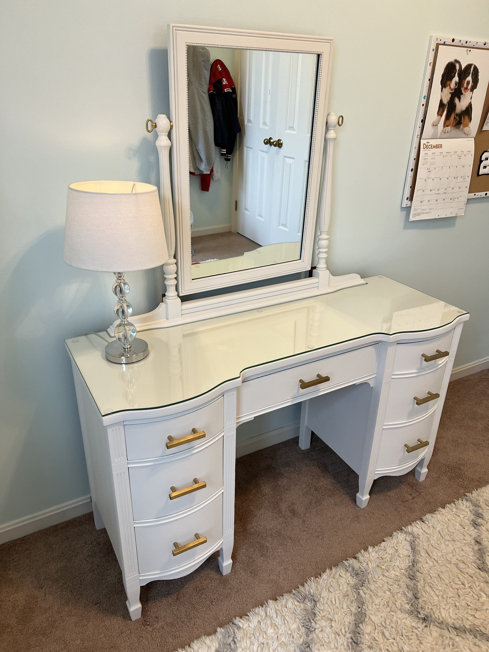 White vanity table with mirror, lamp, and gold drawer pulls against a light blue wall.