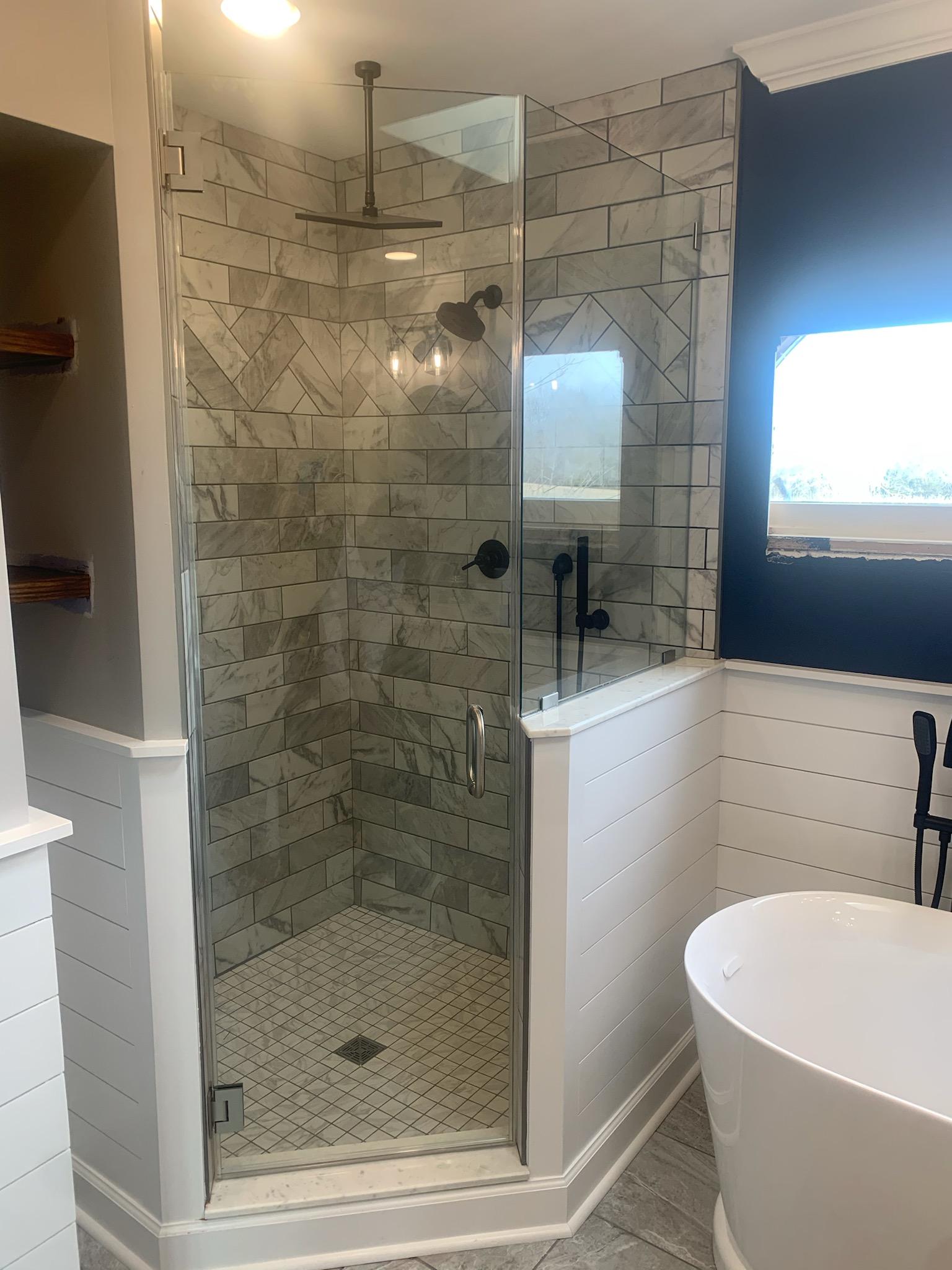 Shower with glass door, herringbone tile, black fixtures. White wainscoting, freestanding tub, and dark blue wall.