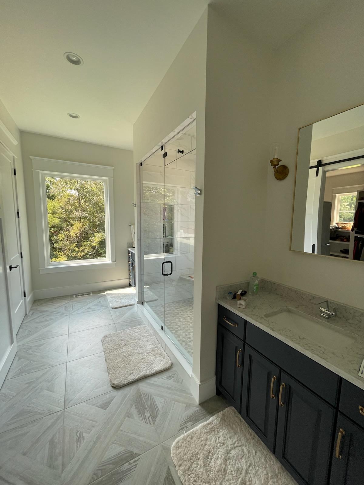 Bathroom with a large window, glass shower, dark blue vanity, and gray tiled floor.