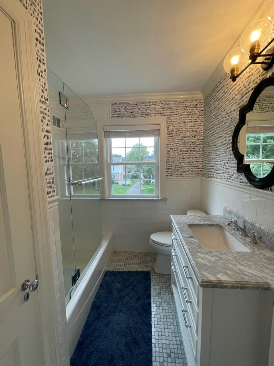 Bathroom with white cabinetry, gray countertop, blue rug, patterned wallpaper, and window.