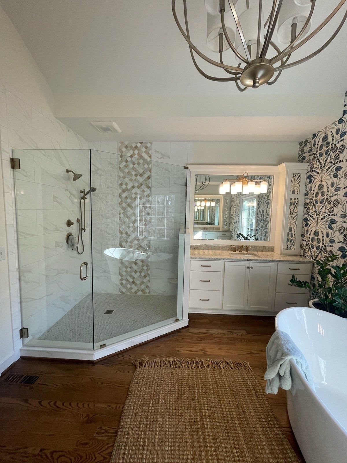 Spacious bathroom with glass shower, white vanity, patterned wallpaper, and hardwood floors.