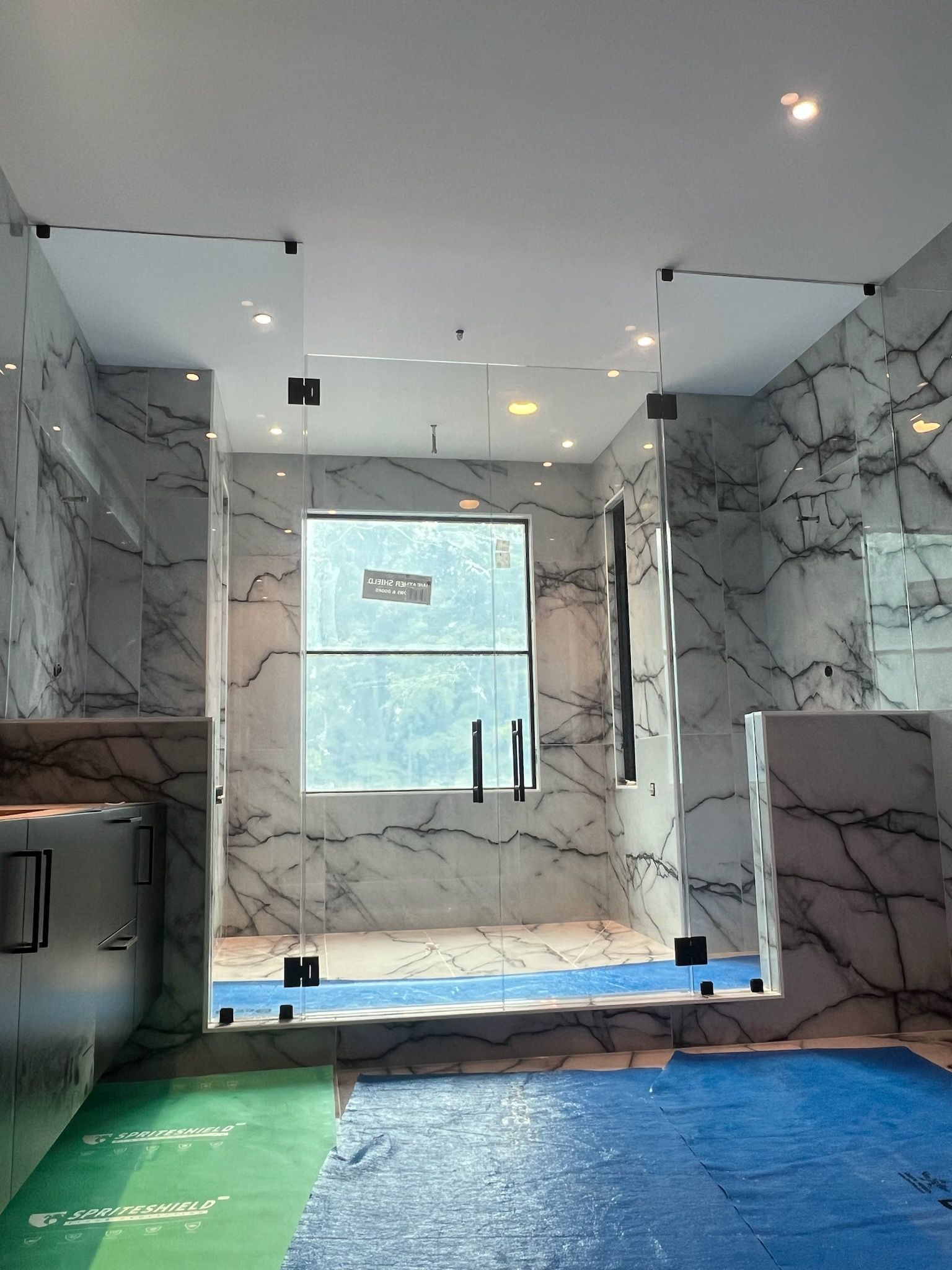 Modern glass shower enclosure with marble walls, black hardware, and blue protective floor covering.