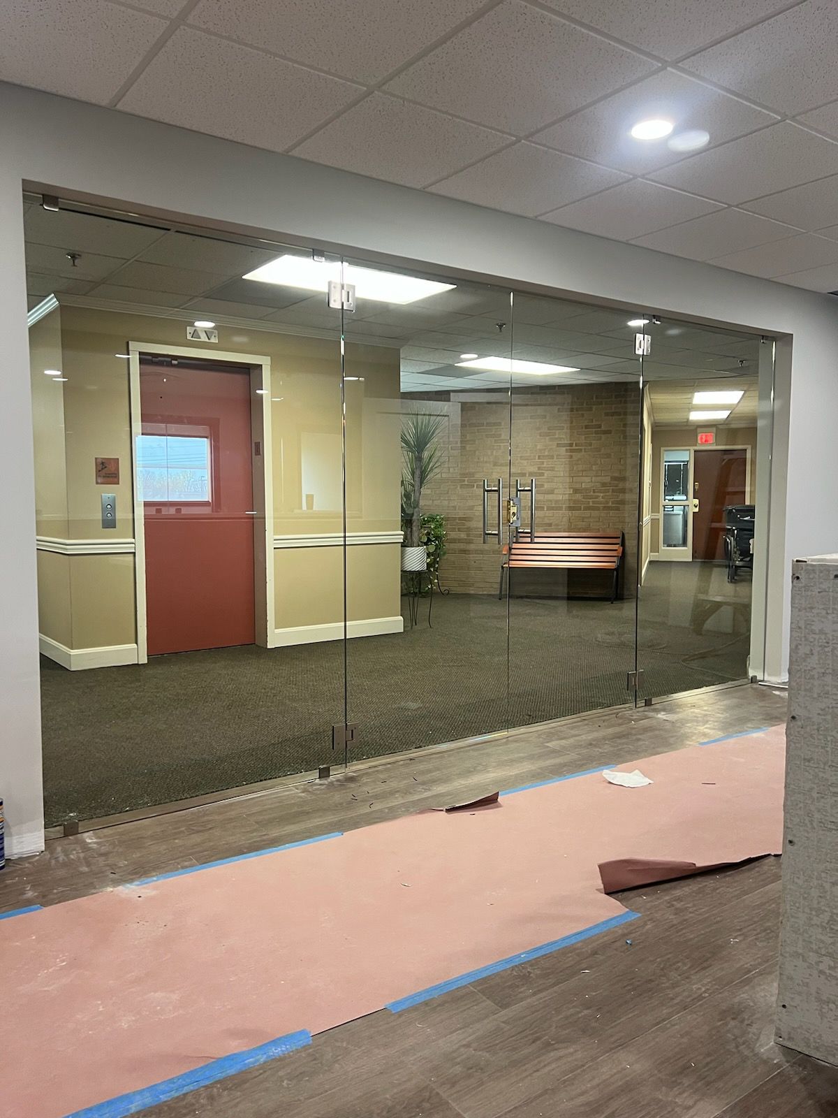 Glass doors open to a hallway with a red door, bench, and carpeting. Construction materials visible.