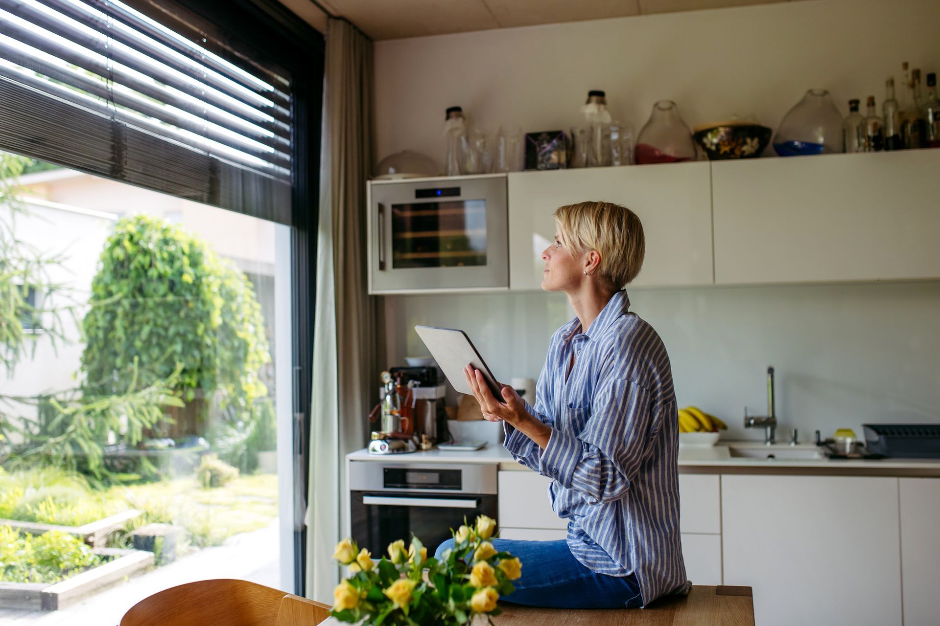 Person holding a tablet while sitting on a kitchen counter near a large window.