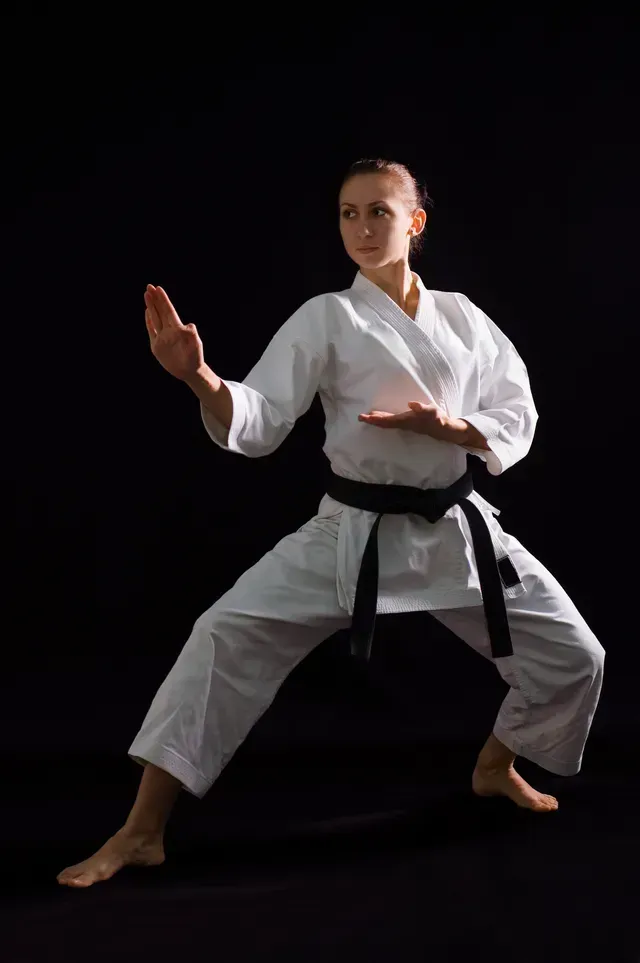 A woman in a white karate uniform with a black belt is practicing karate on a black background.