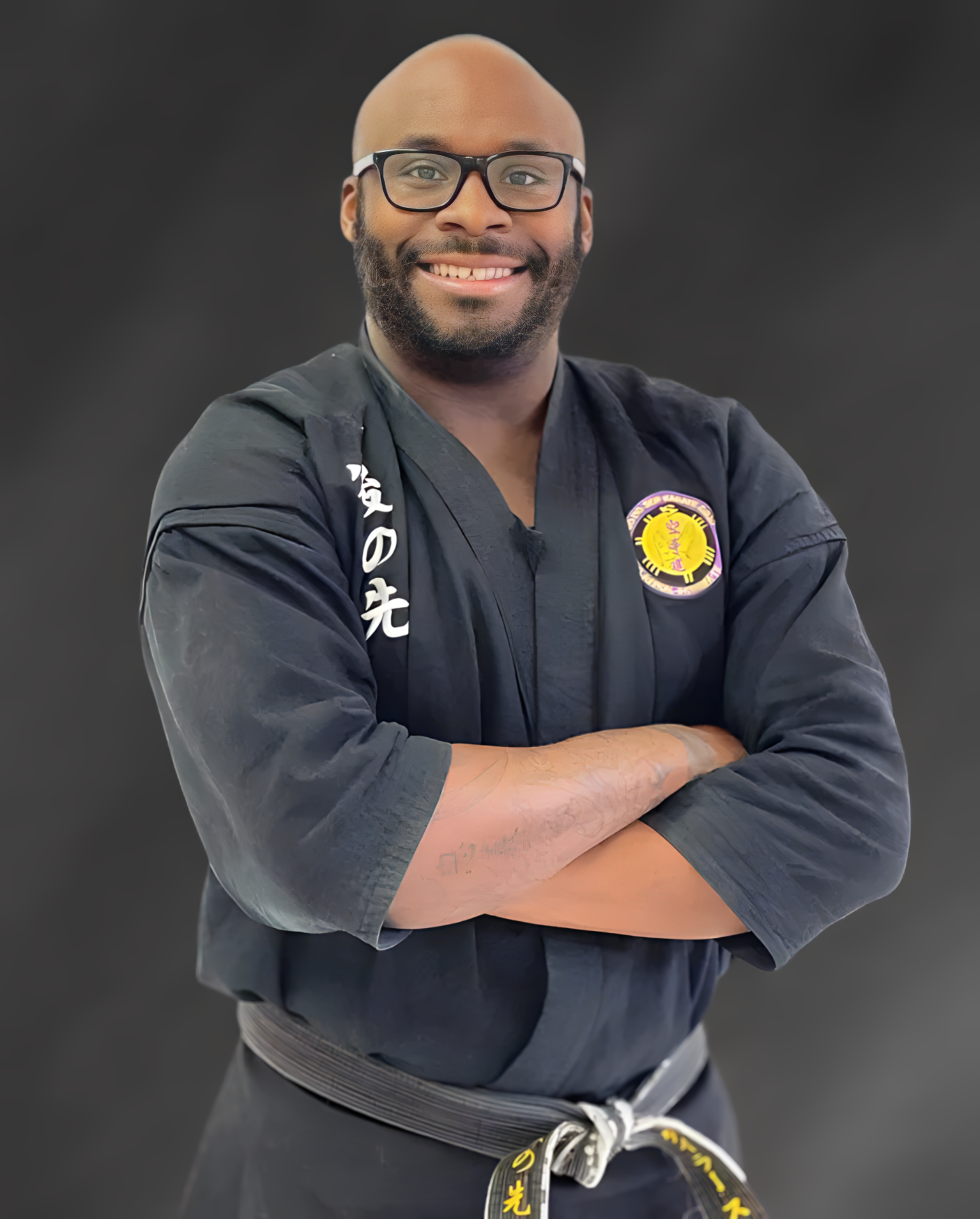 A man in a black karate uniform is smiling with his arms crossed.