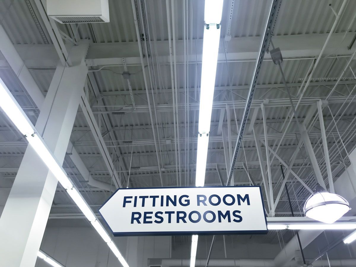 Sign pointing left: Fitting Room, Restrooms. White text on black rectangle. Inside building, overhead lighting.