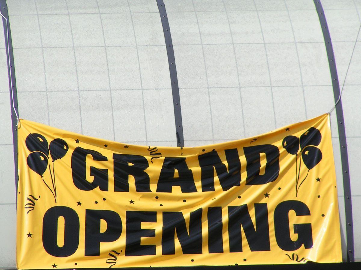 Yellow Grand Opening banner with black text and balloons, hanging in front of a white wall.