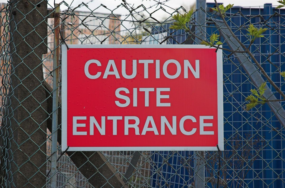 Red CAUTION SITE ENTRANCE sign on a chain link fence in a construction area.