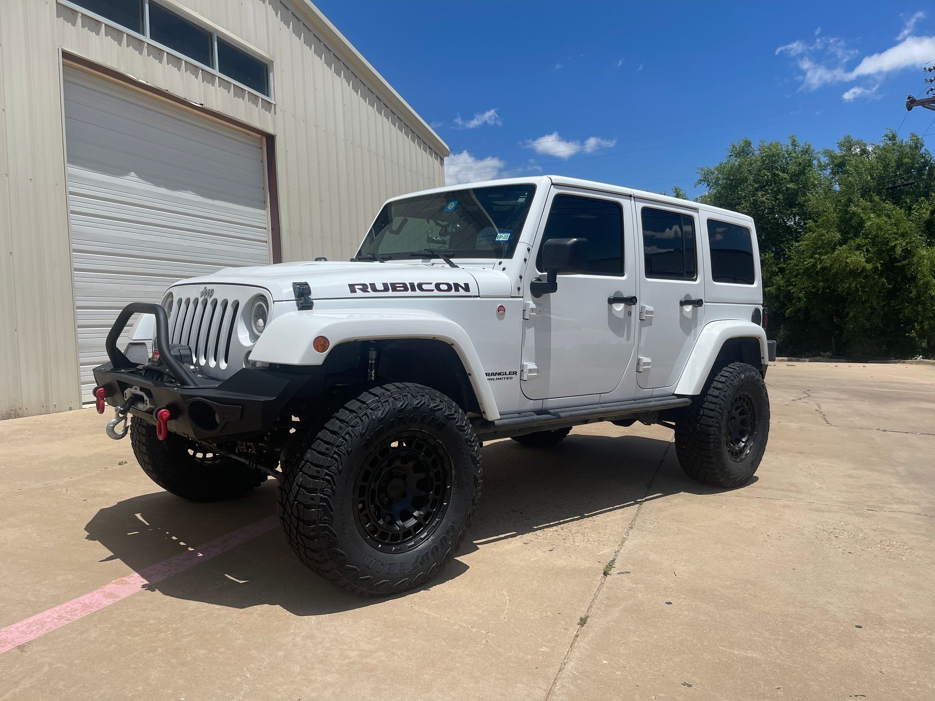 White Jeep Wrangler Rubicon parked in front of a building with black off-road tires and a black bumper.