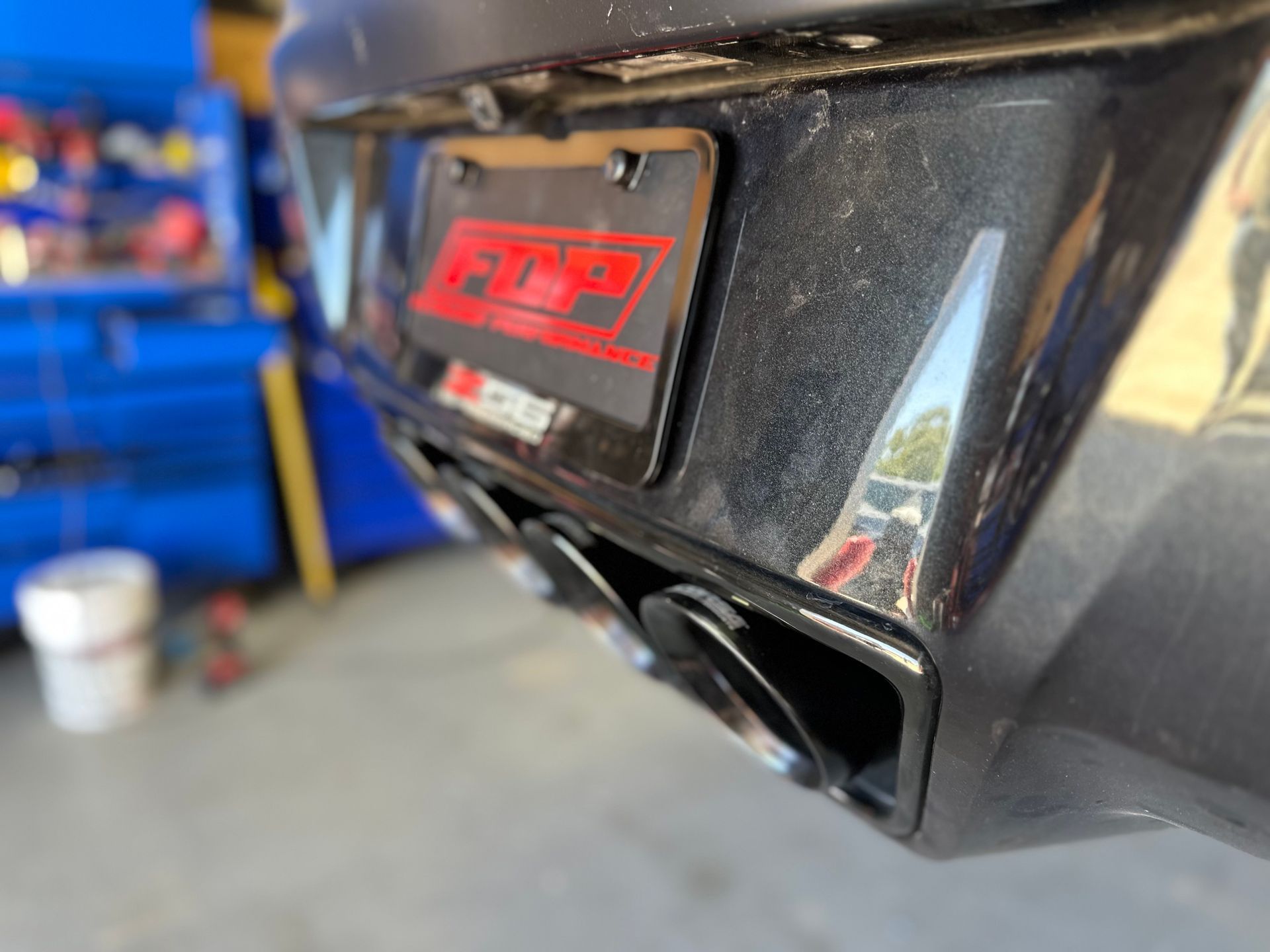 Close-up of a car's black rear bumper with exhaust pipes. A license plate is mounted above.