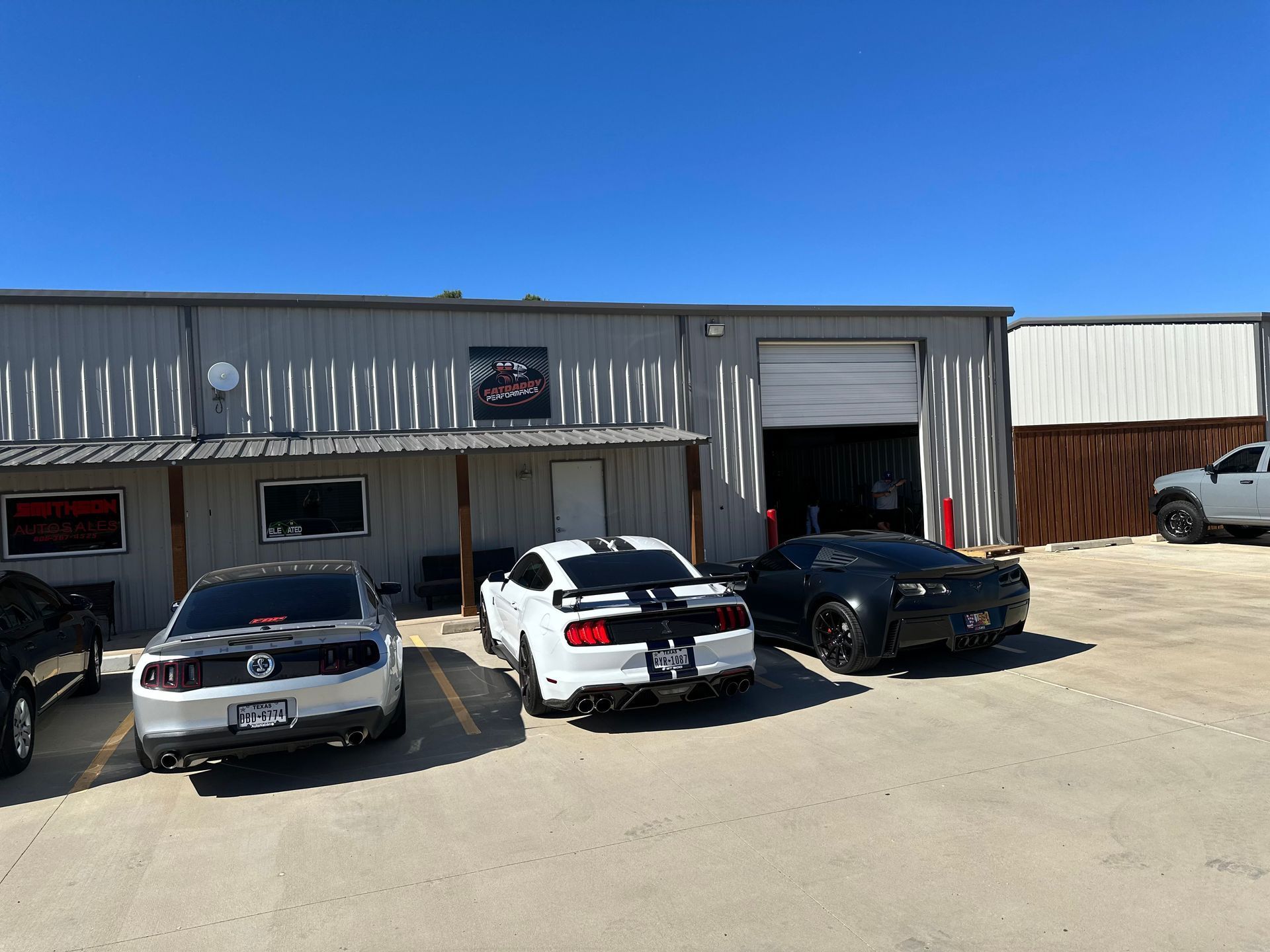 Exterior of a garage with three parked cars: a black, white, and gray Mustang, plus a black Camaro.