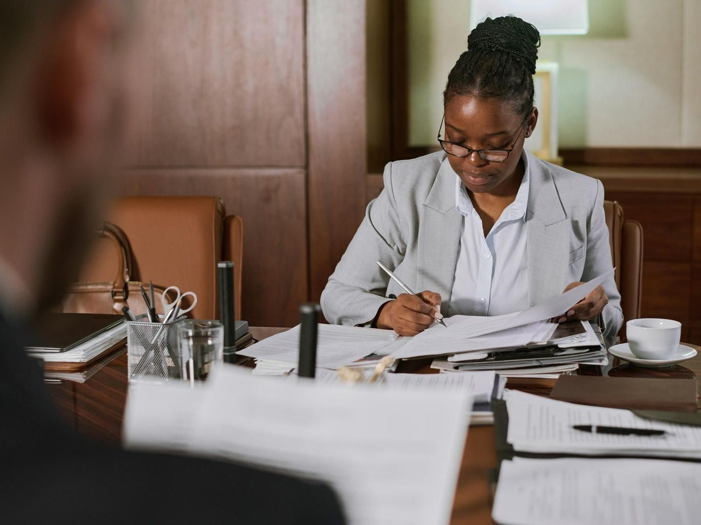 Woman in glasses, gray blazer, writing at a desk, reviewing documents.