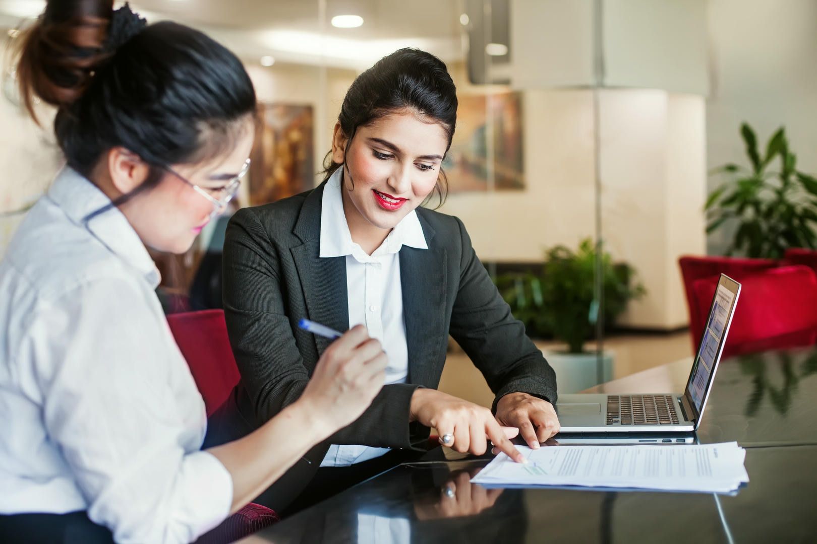 Two women reviewing documents, one points at the paper, and a laptop sits nearby on a table.