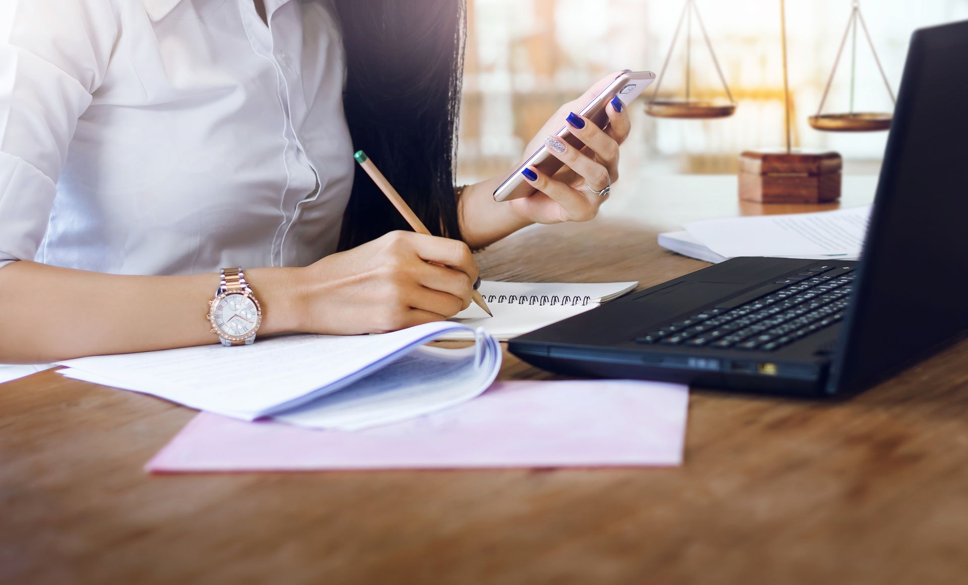 Woman writing with a pencil while using a phone and laptop at a desk with scales of justice.