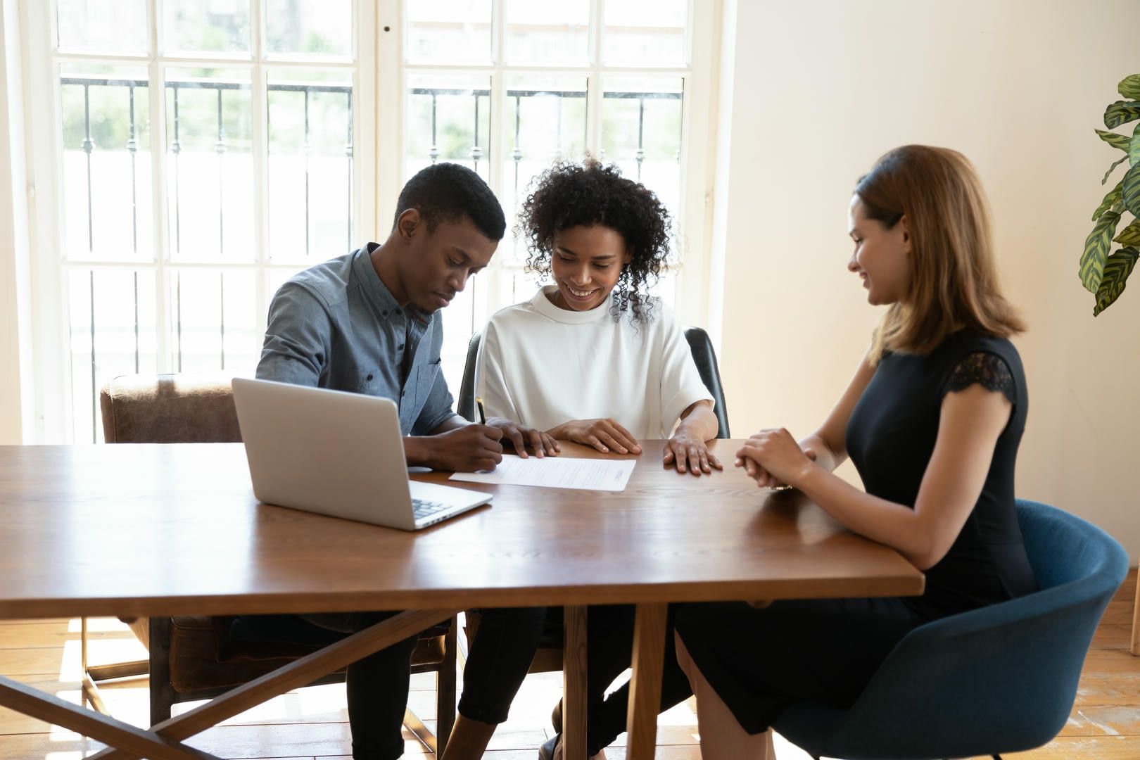 Couple signing documents at a table with a professional. Interior setting with laptop and window.