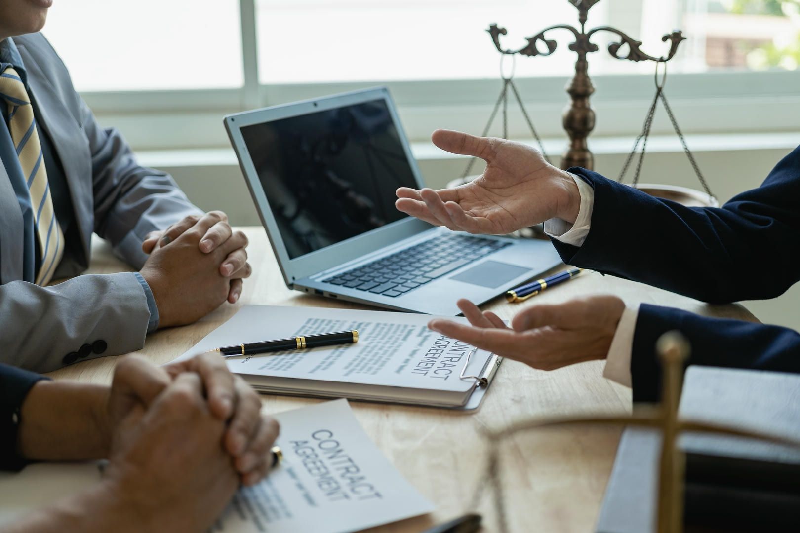 Business meeting with a lawyer; hands gesturing over contract documents, laptop, and scales of justice on a desk.