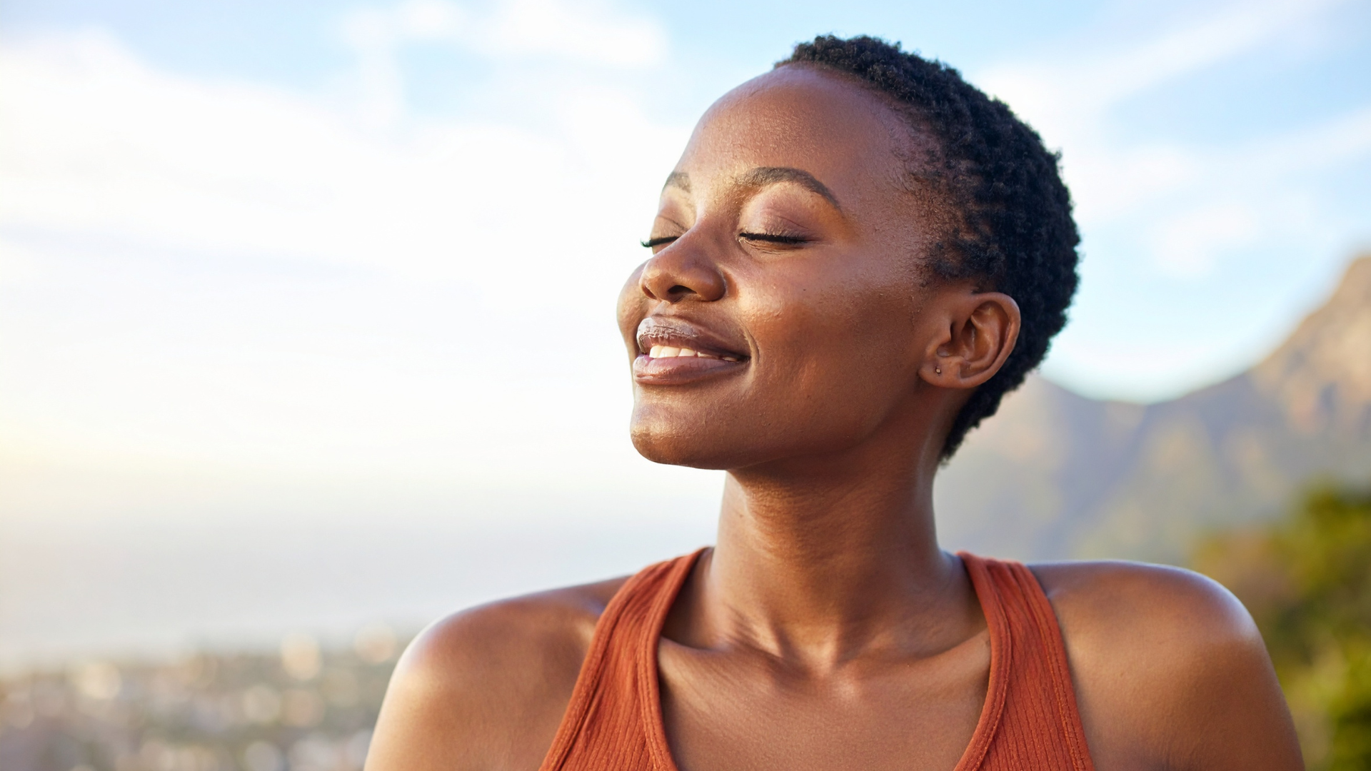 Smiling person outdoors with eyes closed, wearing a rust-colored top, against a bright sky and blurred mountains.
