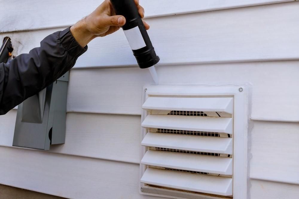 A person is applying sealant to a vent on the side of a house.