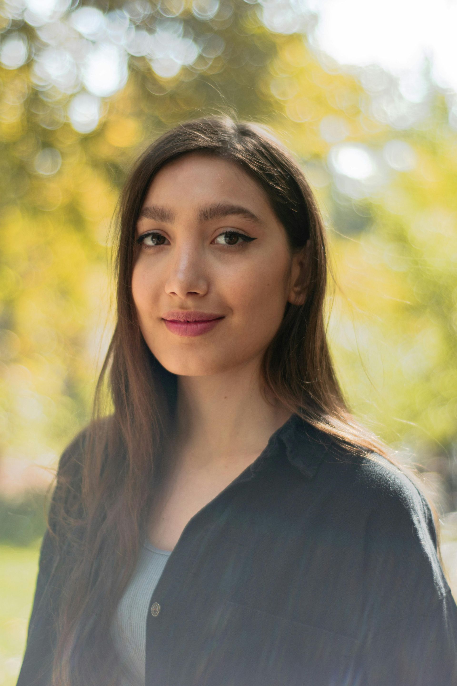 Young woman with long dark hair smiles, wearing a black shirt, against a blurry yellow and green background.